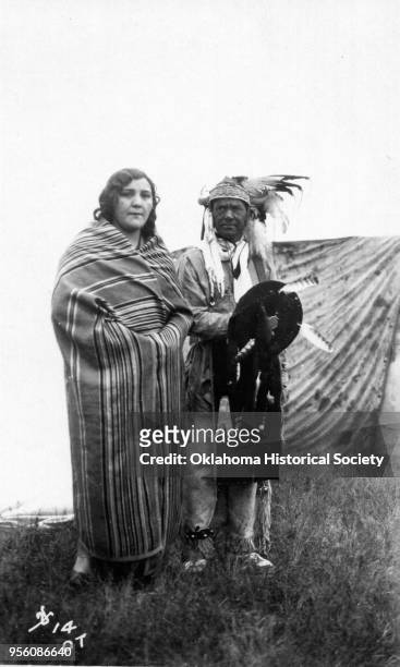 Photograph of an Osage woman wrapped in a wool blanket who is standing with an Osage man wearing a feathered headdress and buckskins, and carrying a...