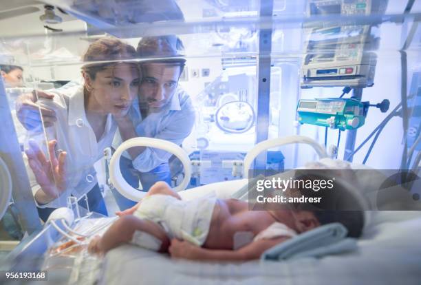 besorgt junges paar betrachten ihre vorzeitigen neugeborenen im brutkasten mit sauerstoff bei neugeborenen intensivstation - family photo in the delivery room stock-fotos und bilder