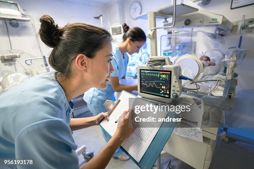 Team of nurses in neonatal intensive care unit checking on a premature's vital signs and taking notes writing the numbers on a clipboard Team of nurses in neonatal intensive care unit checking on a premature's vital signs and taking notes writing the numbers on a clipboard