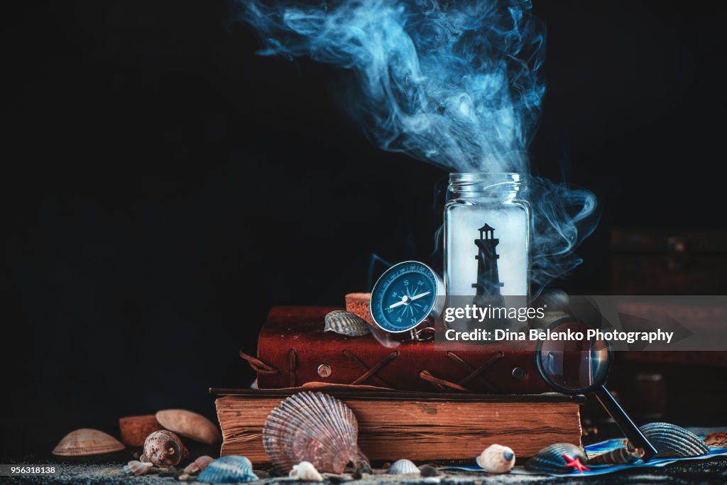 Lighthouse in a glass jar on a stack of books and captain logs. Sea still life with compass, globe and seashells on a dark background with smoke. Marine concept with copy space.