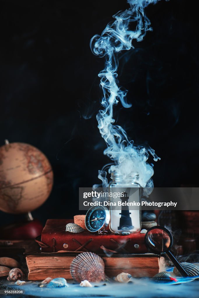 Lighthouse in a glass jar on a stack of books and captain logs. Sea still life with compass, globe and seashells on a dark background with smoke. Marine concept with copy space.