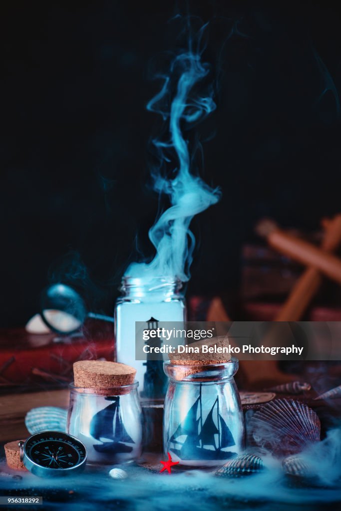 Lighthouse and sailboats silhouettes in glass jars. Sea still life with compass, anchor and seashells on a dark background with smoke. Marine concept with copy space.