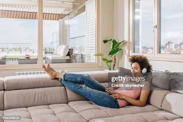 happy black woman using cell phone while listening music over headphones at home. - women of penthouse stock pictures, royalty-free photos & images