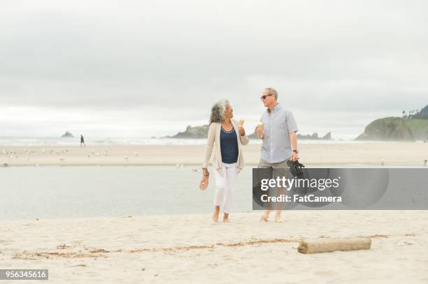 a senior couple walks barefoot along the beach and eats ice cream cones together - oregon coast stock pictures, royalty-free photos & images