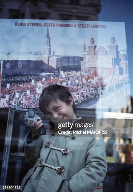 Petit garçon avec un jouet revolver devant une affiche montrant une manifestation pour "le bonheur et le progrès" en novembre 1979 à Prague en...