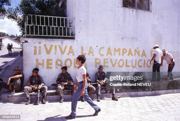 Des militaires assis surveillent la rue devant un mur avec un slogan révolutionnaire, en 1984 au Salvador.