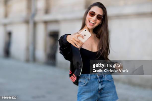 Model Dora Molina taking a photo with her iphone wearing denim shorts and black one piece Urban Outfitters, Quay sunglasses, Just Fabulous boots,...