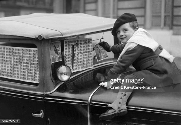 Ce petit garçon colle sur le pare-brise de la voiture des timbres anti-tuberculeux, à Paris, France le 4 décembre 1933.