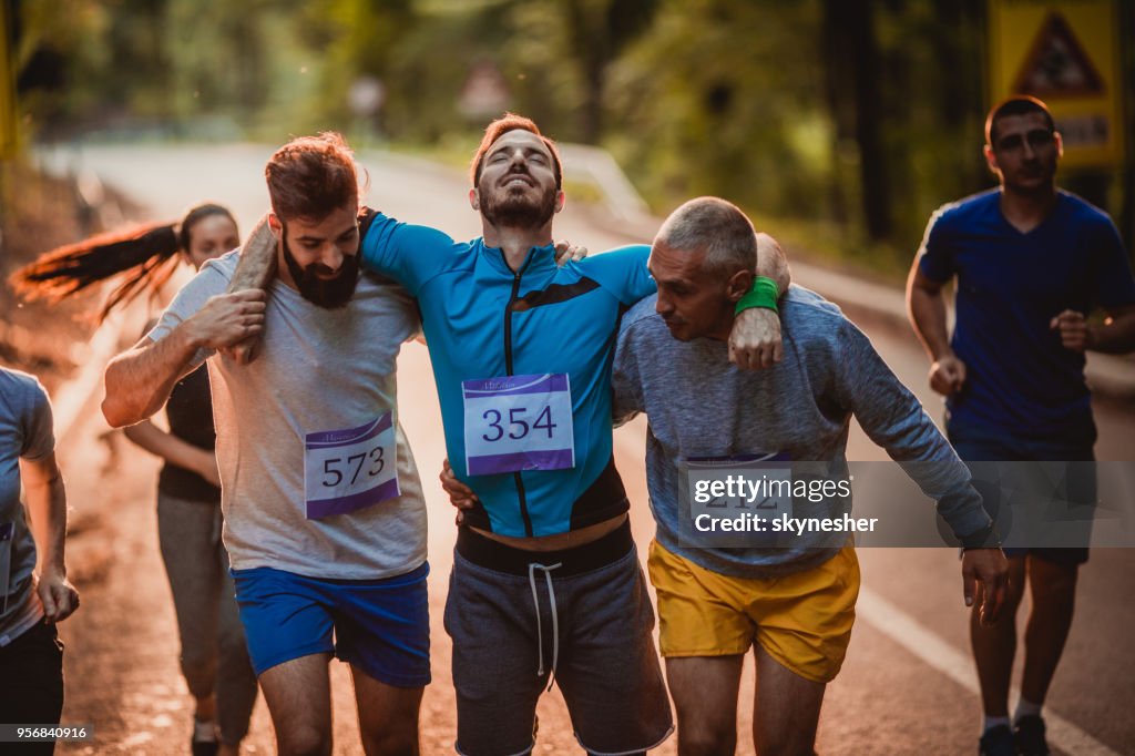 Male runners carrying injured athlete during marathon in nature.