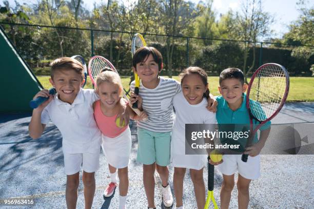 grupo de niños en la cancha de tenis abrazándose uno al otro y sosteniendo sus raquetas mirando a la cámara sonriendo - athletic club fotografías e imágenes de stock