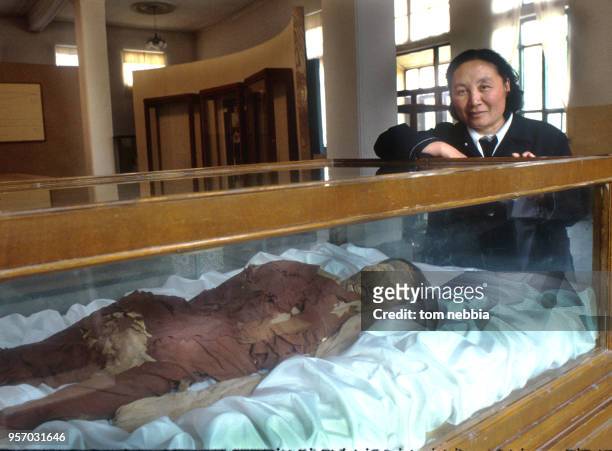 Portrait of a curator at the Urumqi Museum who poses beside a display of mummified corpses in glass cases, Qinghai province, China, April 1980.
