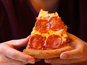 Bitten slice of pizza in the hands of the person close-up. The man in a purple T-shirt with well-groomed hands is eating pepperoni pizza