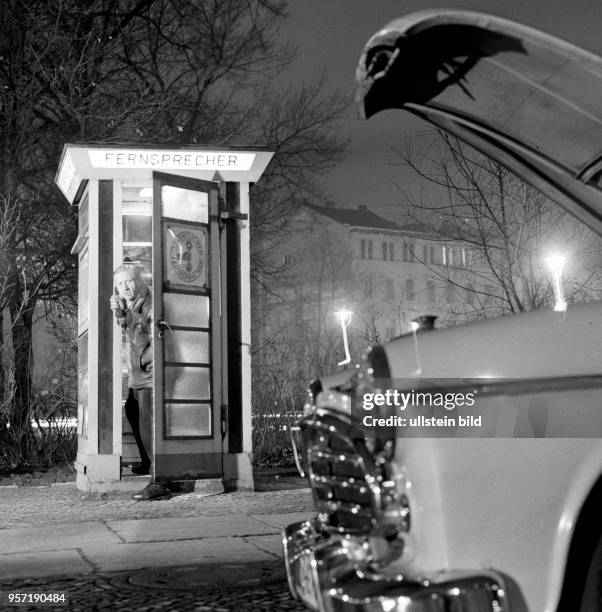 Das offensichtlich gestellte Foto zeigt einen Mann, der aus einer öffentlichen Telefonzelle heraus auf einen mit offener Motorhaube stehenden Pkw...