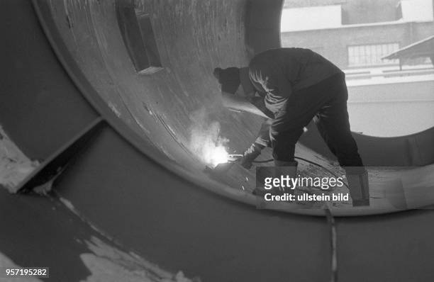 Ein Arbeiter mit Mütze und dicken Filzstiefeln bei Schweißarbeiten an einer Stahlkonstruktion auf einem Montageplatz im Freien im VEB Kranbau...