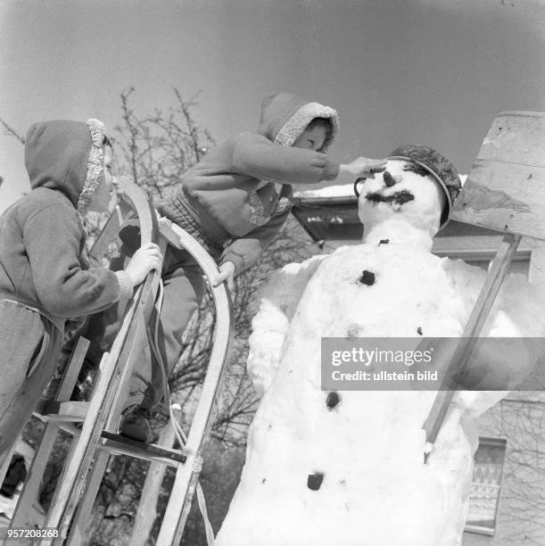 Einen Schlitten als Leiter wird von einem Kind genutzt, um dem großen Schneemann die Möhren-Nase ins Gesicht zu setzen - Kinder bei Spielen...