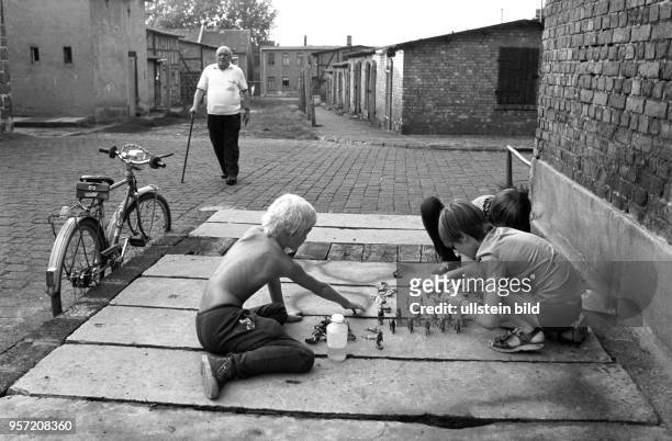 Eine Gruppe Kinder spielt auf der Straße mit Spielzeugfiguren, während ein älterer Mann mit Gehhilfe die Straße überquert, aufgenommen im Frühjahr...