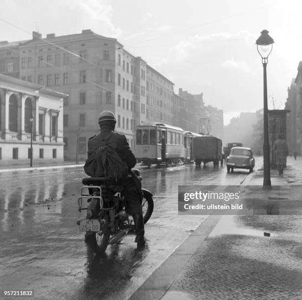 Ein Motorradfahrer mit Helm auf regennasser Fahrbahn in Magdeburg, Straßenverkehr mit Straßenbahn, Pkw und Lkw, aufgenommen 1960.