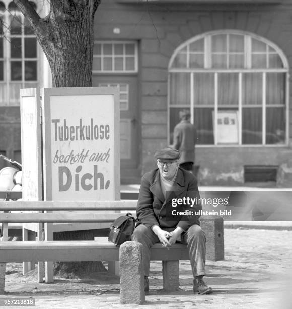 Ein älterer Mann sitzt gähnend auf einer Bank in Schwerin neben einem Aufsteller mit dem Hinweis "Tuberkulose bedroht auch Dich", aufgenommen im...