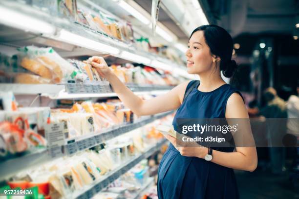 asian pregnant woman grocery shopping in supermarket and choosing packets of cheese - cheese shop stock-fotos und bilder