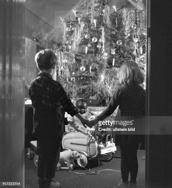 Ein Mädchen und ein Junge, Bruder und Schwester, halten sich an den Händen und betrachten im Wohnzimmer den geschmückten Weihnachtsbaum und die...