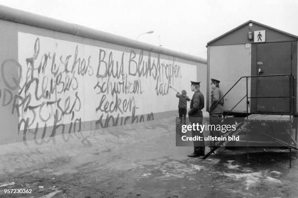 Zwei DDR-Gremzer beobachten am an einem Toilettenwagen stehend, wie ein Teil der Berliner Mauer am Potsdamer Platz von einem Mann mit einem...