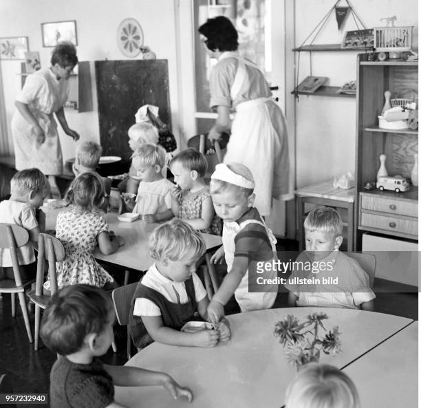 Kinder sitzen an Tischen in einem Kindergarten in Berlin-Treptow, aufgenommen im August 1968. Ein Kind mit Schürze und Haube hilft beim Decken des...