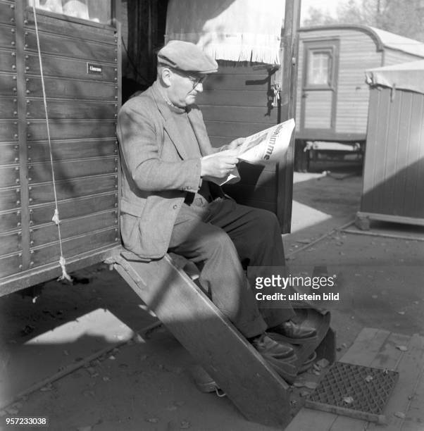 Ein Mann sitzt auf einer Treppe vor enem Holzwagen und liest in einer Tageszeitung , aufgenommen 1963.