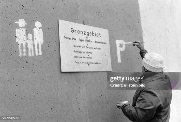 Ein Mann verziert am neben einem Warnschild einen Teil der Berliner Mauer am Potsdamer Platz. Er trägt ein Stirnband mit der Aufschrift "Keine...