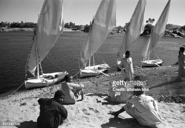 Männer sitzen am Ufer des Nils bei Assuan vor kleinen Booten, sogenannten Feluken, aufgenommen 1972.