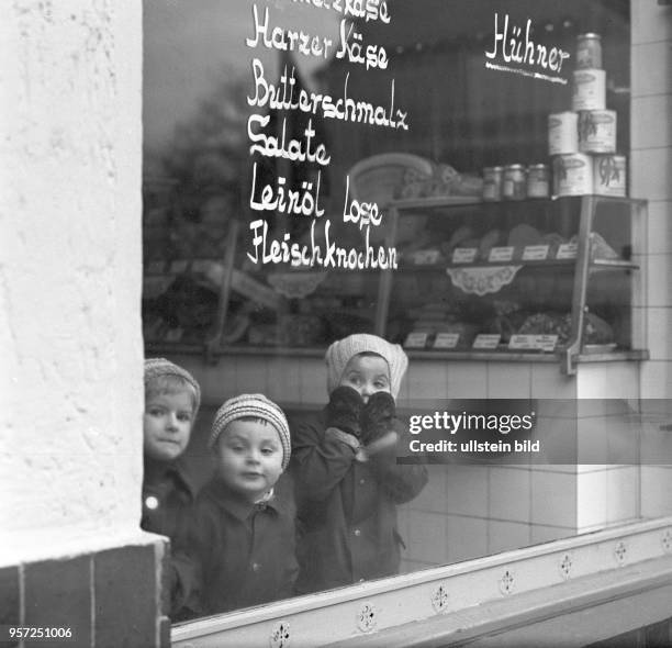 Kinder mit Strickmützen auf den Köpfen stehen in einem kleinen Lebensmittelgeschäft in Berlin und schauen durch die Schaufensterscheibe auf die...