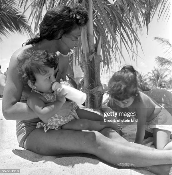 Eine junge Mutter mit ihren Kindern am Strand von Santa Lucia unter einer Palme, , aufgenommen 1962.