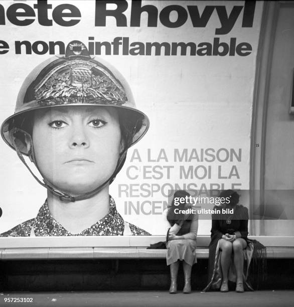 Zwei Frauen sitzen vor einem Werbeplakat für Kunststoff-Textilien und unterhalten sich, aufgenommen im November 1970 in Paris. Die Werbung preist die...