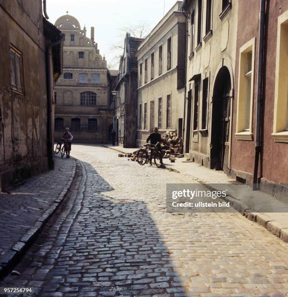 Eine enge Straße mit Kopfsteinpflaster und alte Häuser in der Stadt Güstrow, undatiertes Foto von 1978. Vor einem Wohnhaus liegt ein Haufen Brennholz.