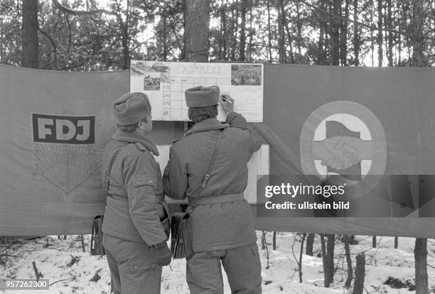 Zwei Offiziere der Nationalen Volksarmee tragen an einer mit dem Logos der FDJ und der SED geschmückten Feldwandzeitung Ergebnisse einer Übung im...