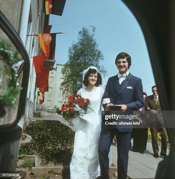 Ein Hochzeitspaar, die Braut mit einem Strauß roter Nelken, einem weißen Brautkleid und einem weißen Schleier, an einer mit DDR-Fahnen geschmückten...