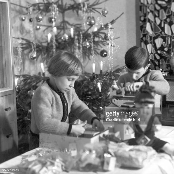 Vor einem mit Glaskugeln, elektrischen Kerzen und Lametta geschmückten Weihnachtsbaum spielen ein Mädchen und ein Junge mit ihren...