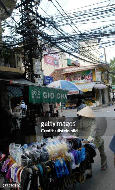 Eine Frau schiebt an einer Straßenkreuzung einen Verkaufsständer mit Schuhen unter einem wahren Gewirr von Stromkabeln in der Altstadt von Hanoi, der...