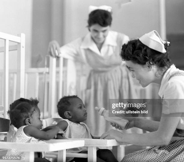 Zwei Frauen als Erzieherinnen geben kleinen Kindern in einem neuen Kindergarten in Santiago de Cuba die Milchflasche, aufgenommen 1962.