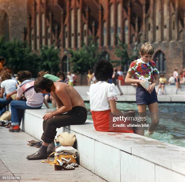 Eine Junge läuft im Wasserbecken der Wasserspiele vor dem Fernsehturm am Berliner Alexanderplatz, Passanten sitzen auf der Mauer, im Hintergrund die...