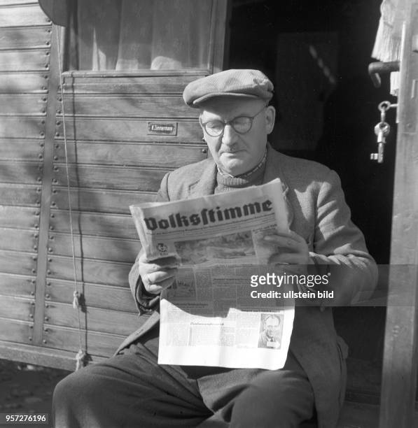Ein Mann sitzt auf einer Treppe vor enem Holzwagen und liest in einer Tageszeitung , aufgenommen 1963.