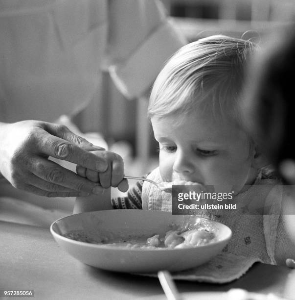 Die Hand einer Erzieherin hilft einem Kind beim Essen an einem Tisch in einem Kindergarten in Berlin-Treptow, aufgenommen im August 1968.
