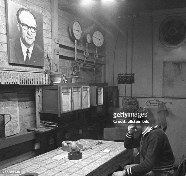 In einem tristen Raum mit Überwachungsschreibern, Manometer und Telefon sitzt ein Kesselwärter unter dem Porträt des DDR-Staatsratsvorsitzenden und...