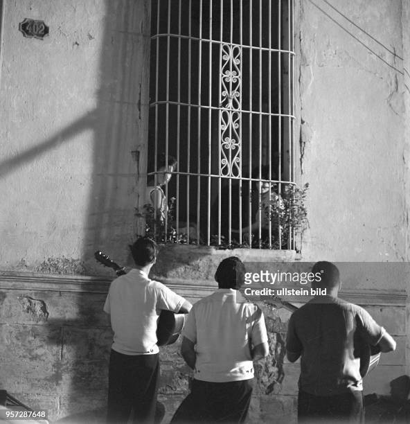 Drei Männer singen an einem vergitterten Fenster, hinter dem zwei Mädchen sitzen, Liebeslieder, aufgenommen in Trinidad 1962.