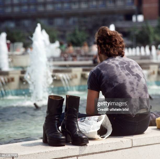 Ein Mann hat seine Stiefel ausgezogen und hält seine Füße in das Wasser der Wasserspiele vor dem Fernsehturm am Berliner Alexanderplatz, .