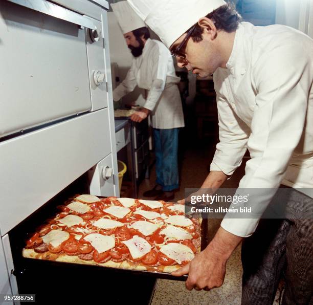 Ein Koch schiebt in der Küche einer Gaststätte ein Backblech in einen Backofen, undatiertes Foto von 1976. Auf dem Blech lieg ein mit Wurst-,...