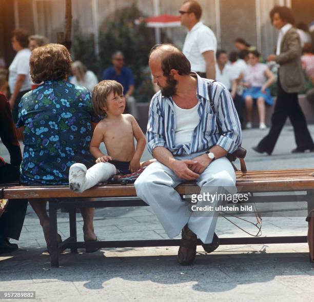 Ein Mann sitzt neben einem Jungen mit Gipsbein auf einer Bank am Alexanderplatz in Berlin, aufgenommen am .