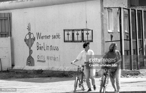 Zwei Frauen mit Fahrrädern vor einer Zeichnung der Comic-Figur "Werner" und der Schriftzug "Werner sacht: Bärlin muss oins werden" an der Berliner...