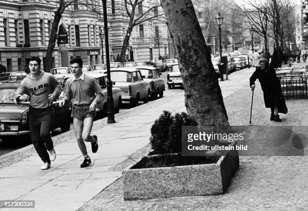 Junge Männer beim Joggen in der Husemannstraße in Berlin-Prenzlauer Berg, aufgenommen 1988. Eine alte Dame mit Gehhilfe blickt den beiden...