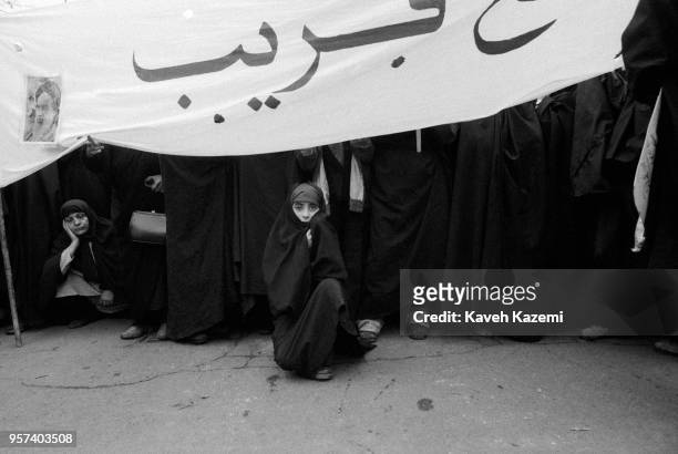 Women demonstrators, among them a young girl covered in black chador sits under a banner with a photo of Ayatollah Khomeini in the front row during a...