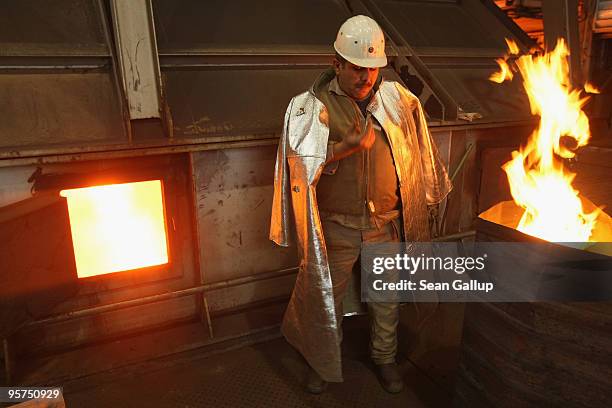 Worker wearing protective clothing takes a cigarette break as molten iron from a blast furnace flows into a container at left while a fire burns to...